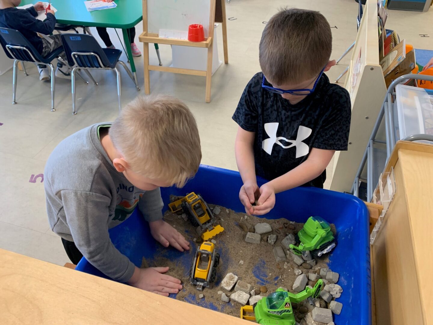 Children playing with toy trucks in sand.