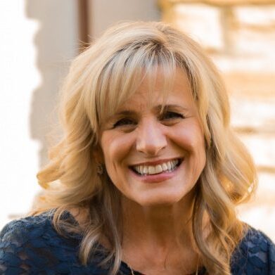 A smiling blonde woman with curly hair in a blue top.