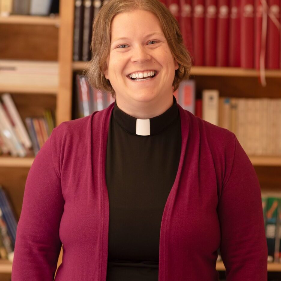 A smiling clergy woman in a maroon cardigan and clerical collar.
